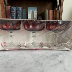 May include: A set of three ruby red stemmed glasses in a cardboard box, wrapped in clear plastic. The glasses have a faceted bowl design and clear glass stems. The box is white with pink and red accents.