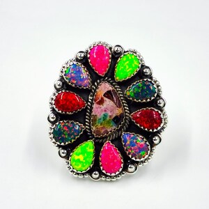 Puede incluir: Un anillo de plata con una piedra central multicolor rodeada por un halo de gemas en forma de lágrima en tonos rojo, rosa, verde y azul. El anillo tiene una banda de plata detallada y texturizada.