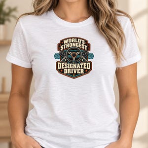 May include: A white short-sleeve t-shirt featuring a graphic design on the chest. The graphic includes a steering wheel, two car keys, and the text "WORLD'S STRONGEST DESIGNATED DRIVER" in a bold, multi-colored emblem with orange and blue accents.