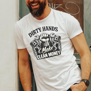 Może przedstawiać: Biała koszulka z napisem "DIRTY HANDS MAKE CLEAN MONEY" w czarno-białej grafice. Grafika zawiera dłoń trzymającą klucze i tłoki. Koszulka jest noszona przez osobę.