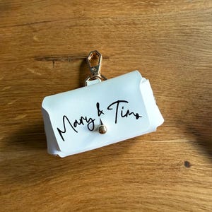 May include: A white leather pouch with a gold-colored clasp and a small gold-colored button closure. The words "Mary & Tina" are written in black script on the front of the pouch. The pouch is designed to hold small items.