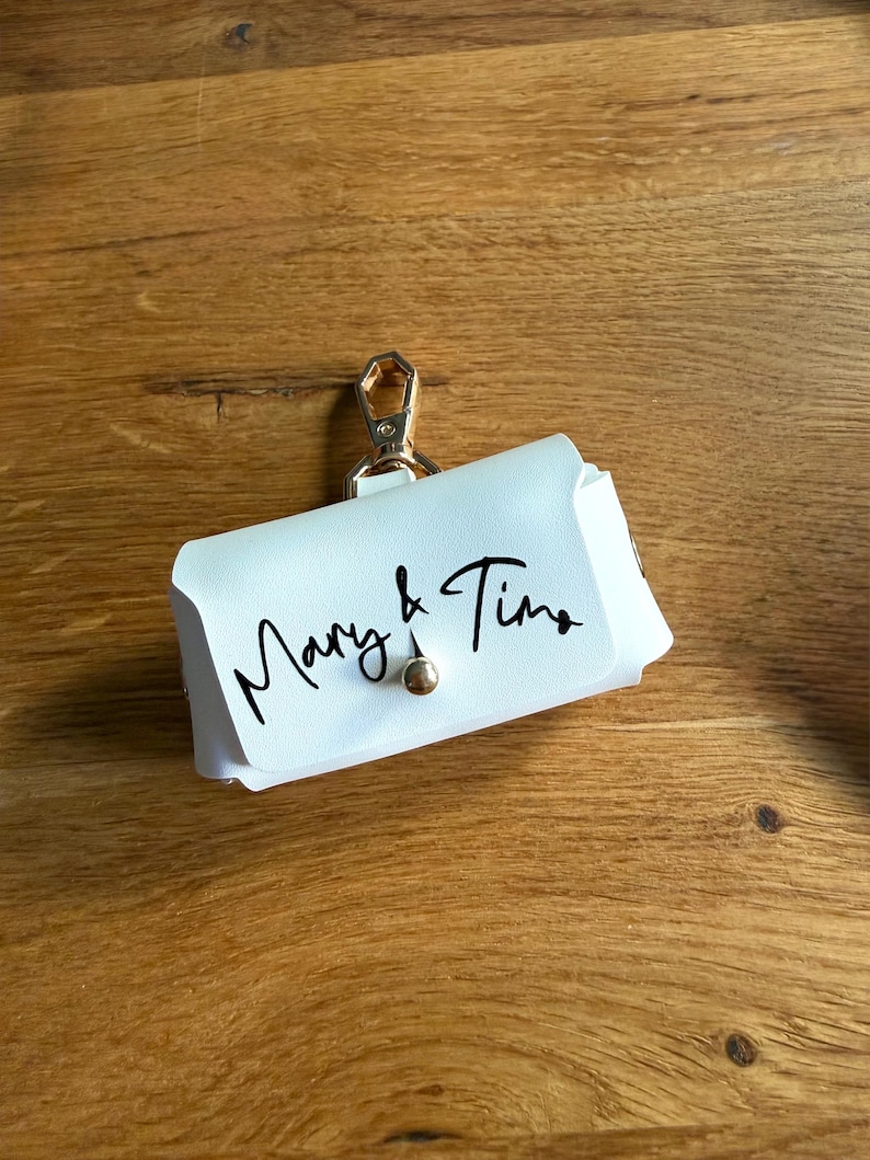 May include: A white leather pouch with a gold-colored clasp and a small gold-colored button closure. The words "Mary & Tina" are written in black script on the front of the pouch. The pouch is designed to hold small items.