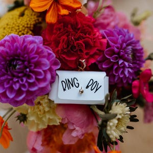 May include: A vibrant bouquet of flowers in shades of purple, red, orange, and pink. A small white card with the words "RING BOY" is nestled among the blooms. A gold ring is visible.
