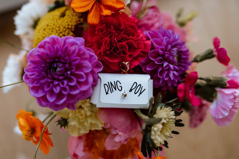 May include: A vibrant bouquet of flowers in shades of purple, red, orange, and pink. A small white card with the words "RING BOY" is nestled among the blooms. A gold ring is visible.