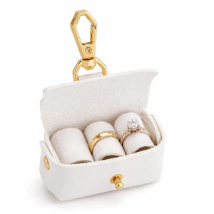 May include: White leather ring box with a gold-tone clasp and hardware. The interior holds three ring rolls, displaying a gold band and a diamond engagement ring. A stylish accessory for jewelry storage.