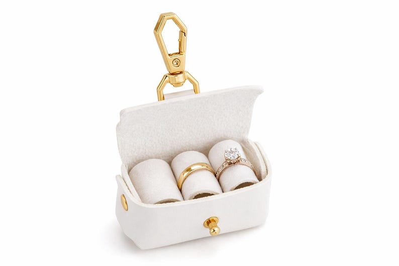 May include: White leather ring box with a gold-tone clasp and hardware. The interior holds three ring rolls, displaying a gold band and a diamond engagement ring. A stylish accessory for jewelry storage.