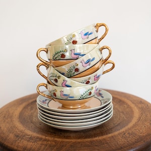 May include: A stack of vintage teacups and saucers. The cups are cream-coloured with gold handles and feature a painted design of pink birds, blue wings, and red berries. The saucers are stacked below the cups.