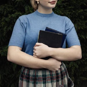 May include: A light blue short-sleeved sweater with a textured pattern around the neckline. The person is wearing a plaid skirt and holding two books. The books are black and blue. The background is a dark green.