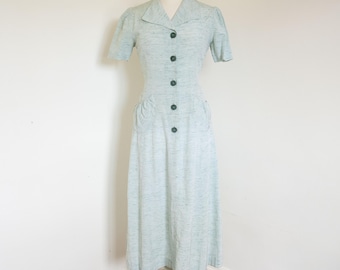 May include: A light blue vintage dress with short sleeves and a collared neckline. The dress features a button-down front, pockets, and a midi-length skirt. The dress is displayed on a mannequin.