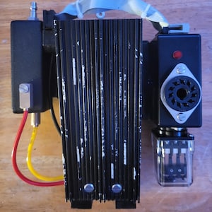 May include: A black electronic component with a ribbed, heat-sink-like structure. It has various connectors, a red light, and a clear plastic component. The unit is mounted on a wooden surface and has a white cable.