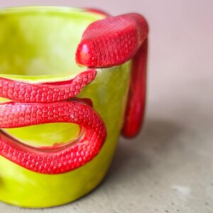 Puede incluir: Una taza de cerámica con un exterior amarillo verdoso brillante y una serpiente roja enrollada alrededor del lateral. Las escamas de la serpiente son detalladas y la taza tiene un acabado liso y brillante. La taza es una pieza única, hecha a mano.