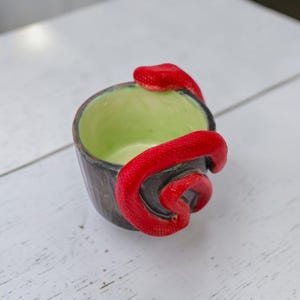 Op de afbeelding: Een kleine, handgemaakte keramische kom met een opgerolde rode slang rond de buitenkant. De kom heeft een donkere, getextureerde buitenkant en een lichtgroene binnenkant. De slang is felrood.