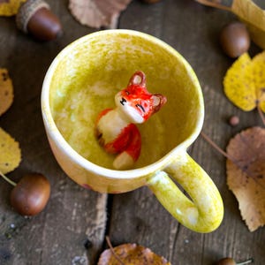 Puede incluir: Una taza de cerámica amarilla con una figura de zorro rojo y blanco en su interior. La taza tiene un asa curva y una superficie texturizada. Bellotas y hojas de otoño rodean la taza sobre una superficie de madera.