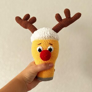 May include: A crocheted reindeer made to look like a beer mug. The mug is yellow with a white foam top and has a red nose and black eyes. The reindeer has brown antlers.
