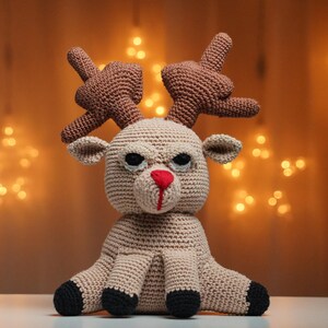 Könnte beinhalten: Ein gehäkeltes Rentier-Spielzeug mit braunen Geweihen, einem beigefarbenen Körper und schwarzen Augen. Das Rentier hat eine rote Nase und sitzt auf einer weißen Oberfläche.