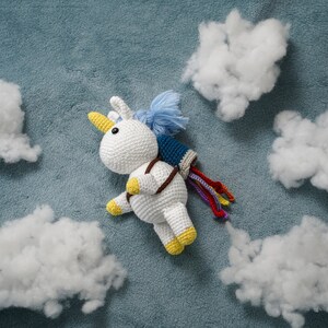 Puede incluir: Un juguete de unicornio blanco de crochet con pezuñas amarillas y un paquete de cohetes de crochet azul y blanco volando a través de nubes de algodón blanco sobre un fondo azul.