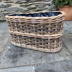 May include: A rectangular wicker basket planter with a woven design. The basket is made of light brown and grey wicker and has a black interior lining. The planter is designed for outdoor use and is suitable for flowers or plants.