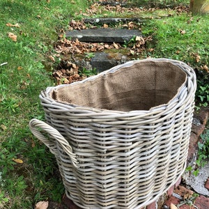 May include: A large, grey wicker basket with a burlap lining sits on a brick wall. The basket has a curved handle. Green grass and a stone staircase are in the background. The basket is suitable for storage or decorative use.