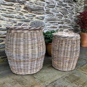 May include: Two woven wicker storage baskets with lids. The larger basket is cylindrical and the smaller one is more oval. Both are light brown with a white wash finish. The baskets are on a stone patio.