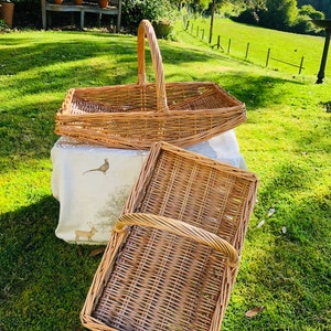 May include: Two light brown woven wicker baskets with handles. One is rectangular with a tall handle, the other is a rectangular tray with a shorter handle. They rest on a white cloth with a pheasant and deer pattern.