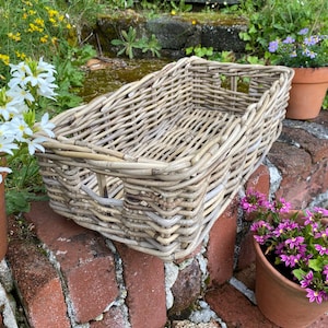 May include: A rectangular, light grey wicker basket with cut-out handles. The basket is set on a brick surface, with potted flowers and greenery in the background. The basket is empty and ready to be filled.