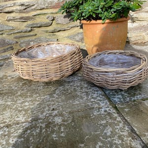 May include: Two round, woven wicker baskets with plastic liners. A terracotta pot with green plants is in the background. The baskets are light brown and sit on a stone surface. The scene is outdoors.