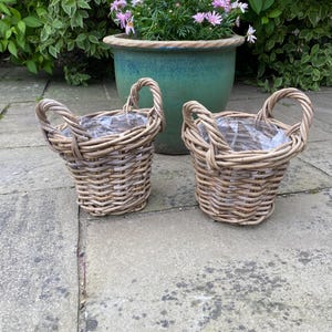 May include: Two small, woven wicker baskets with handles, ideal for planting. The baskets are light brown and sit on a stone patio. A large, turquoise ceramic pot with pink flowers is in the background, surrounded by green foliage.