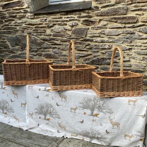 May include: Three rectangular wicker baskets with arched handles. The baskets are displayed on a table covered with a white fabric featuring a woodland scene with deer, trees, and birds. The baskets are various sizes.