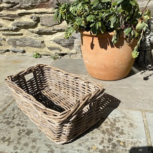 May include: A rectangular wicker basket in light brown sits on a stone patio. The basket is empty and has a woven design. A large terracotta pot with a green plant is in the background. A stone wall is visible.