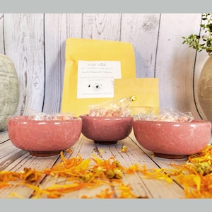 May include: Three pink ceramic bowls with a crackle glaze, filled with small, wrapped candies. A yellow package with text and a small packet are behind the bowls. Dried orange flowers are scattered on the wooden surface.