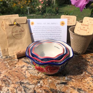 Puede incluir: Un juego de tres cuencos medidores de cerámica con un estampado floral, ribete rojo y azul, y una marca de 60 ml. Los cuencos están anidados. La imagen también incluye sobres de semillas, un corcho y una tarjeta con texto sobre polinizadores.
