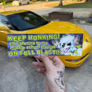 以下が含まれることがあります： 「KEEP HONKING! im listening to my middle school playlist ON FULL BLAST!!!」の文字が入ったバンパーステッカーが、黄色のスポーツカーの前で掲げられています。ステッカーは紫と緑の背景に漫画のキャラクターが描かれています。
