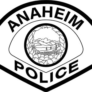 May include: Black and white badge with the words "ANAHEIM POLICE" in a circular design. The badge features a stylized image of a landscape with a small town and trees.