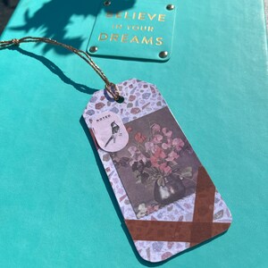 以下が含まれることがあります： 「Believe in your dreams」と書いてあるターコイズブルーのノート。ノートには、花柄と鳥のイラストが描かれた茶色と白のしおりが付けられています。