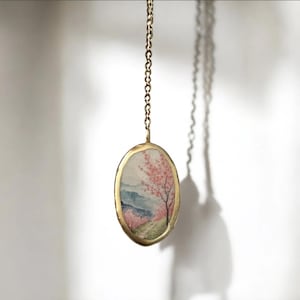 May include: A gold chain necklace with an oval pendant featuring a watercolor painting of a pink tree and a landscape. The pendant has a textured gold rim.