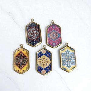 May include: Five decorative pendants with a gold-colored frame and a small loop at the top. Each pendant features a miniature rug design in various colors, including blue, pink, yellow, and white. The pendants are displayed on a white marble surface.