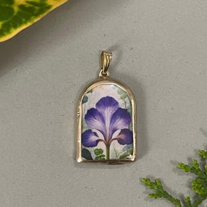 Peut inclure: Pendentif doré représentant une fleur d'iris violette peinte à la main. Le pendentif a une forme arquée et une bélière pour une chaîne. L'iris est placé sur un fond de feuillage vert et un arrière-plan clair.
