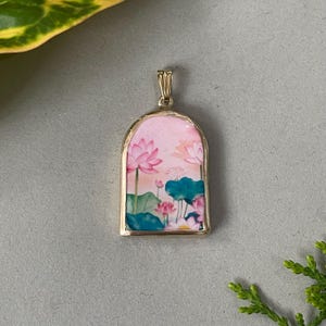 May include: A gold-colored pendant with a rectangular shape and a rounded top. The pendant features a watercolor painting of pink lotus flowers, green leaves, and blue-green lily pads against a pink background.