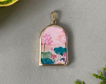 May include: A gold-colored pendant with a rectangular shape and a rounded top. The pendant features a watercolor painting of pink lotus flowers, green leaves, and blue-green lily pads against a pink background.