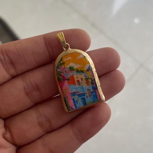 May include: Gold-toned pendant with a colorful painted scene of a coastal town. The pendant features a rectangular shape with rounded top edges. The artwork depicts buildings in shades of blue, pink, and orange, with palm trees and a sunset sky.