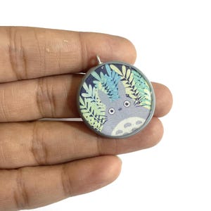 May include: A round pendant featuring a cartoon character, surrounded by green and blue foliage against a dark blue background. The pendant has a silver-colored metal frame and a small loop at the top for a chain.