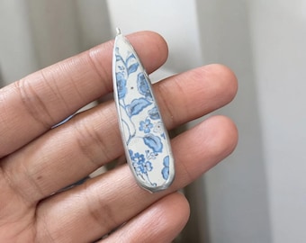 May include: A teardrop-shaped pendant with a white background and a blue floral pattern. The pendant is made of metal and has a silver finish.