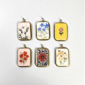 Puede incluir: Seis colgantes rectangulares con diseños florales. Cada colgante tiene un marco dorado y un pequeño lazo en la parte superior. Los diseños florales incluyen margaritas, amapolas y otras flores coloridas sobre varios fondos.