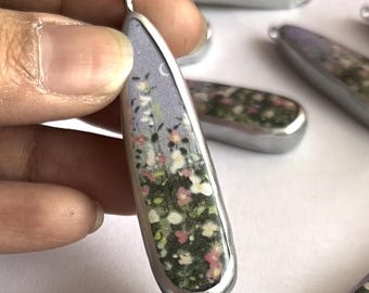 May include: A silver pendant with a floral design inside. The pendant is shaped like a teardrop and has a silver border. The floral design is a mix of pink, white, and green flowers on a light purple background.