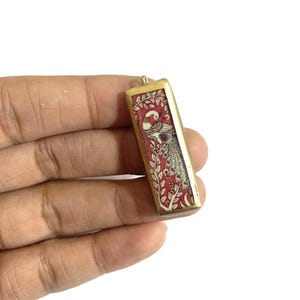 May include: Rectangular pendant with a gold-colored frame. The pendant features a red background with a detailed illustration of a peacock and foliage. The peacock is depicted in white and gray with red accents.