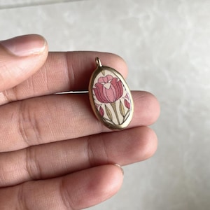 May include: Oval pendant with a floral design. The pendant features a pink and red tulip design on a cream background, set within a gold-colored metal frame. The pendant is small, approximately 1 inch in length.