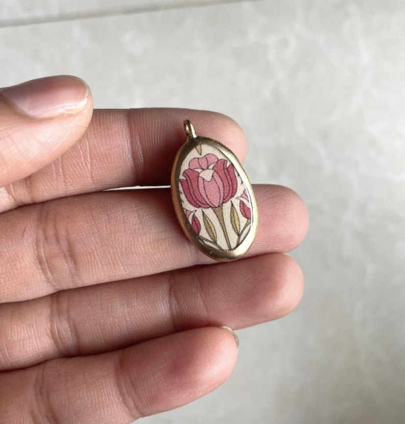 May include: Oval pendant with a floral design. The pendant features a pink and red tulip design on a cream background, set within a gold-colored metal frame. The pendant is small, approximately 1 inch in length.
