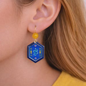 Puede incluir: Un pendiente dorado con una cuenta amarilla y un colgante hexagonal azul. El colgante presenta un diseño de serpiente blanca, formas de media luna y un borde dorado. El pendiente se lleva en la oreja de una persona.