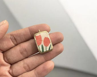 May include: A gold-toned pendant with a rectangular shape features a floral design of three red tulips with green stems and leaves on a white background.