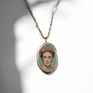 Könnte beinhalten: Eine goldene ovale Halskette mit einem Anhänger, der ein Porträt von Frida Kahlo zeigt. Der Anhänger hat einen blauen Hintergrund und eine rosa Blumenkrone.